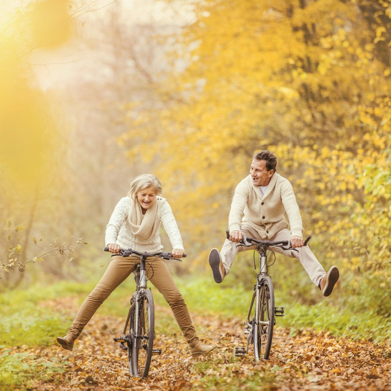 Couple joyfully riding bicycles on a path covered in fallen leaves, autumn setting.