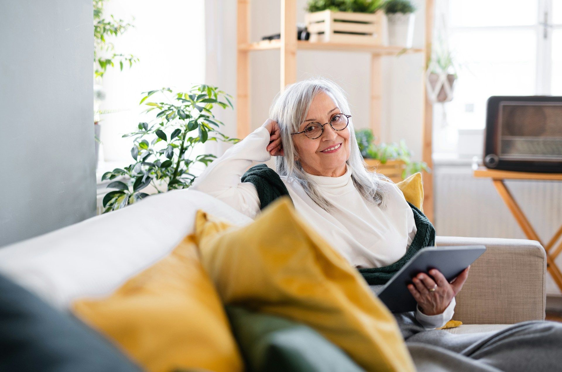 Woman with gray hair smiles while using a tablet on a couch; indoor setting with plants and shelves.