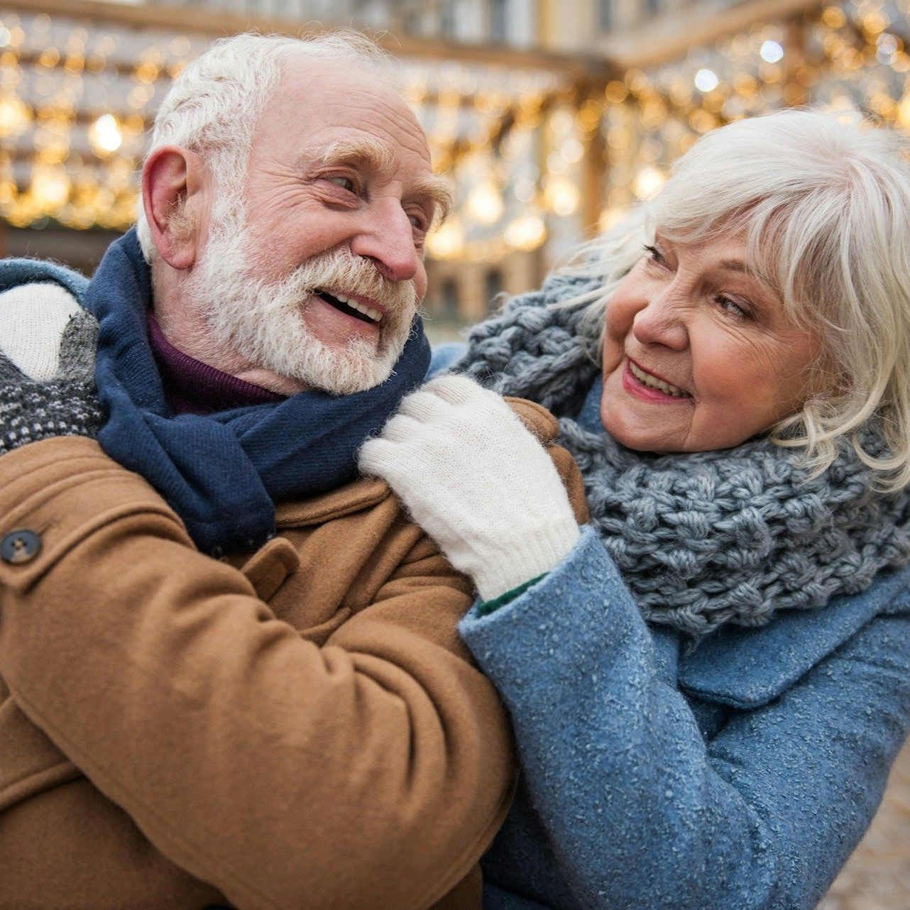 Elderly couple laughing, embracing outdoors, winter clothing. Background: string lights.