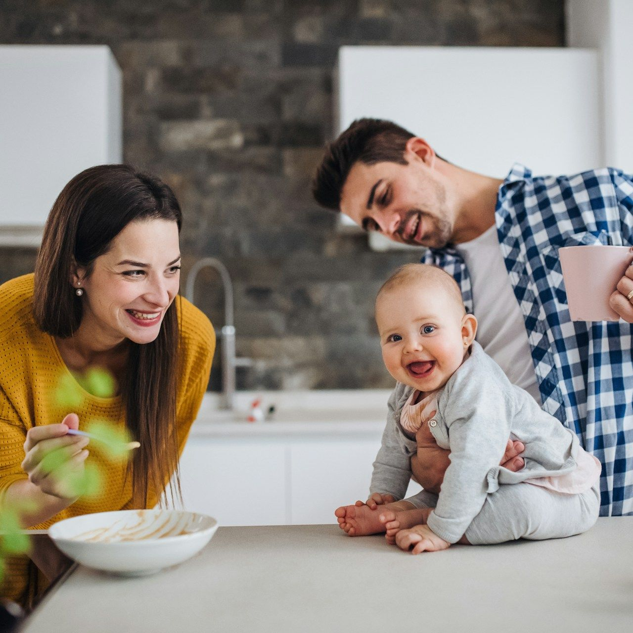 A family in a modern kitchen. Parents smile at baby sitting on counter, near a bowl of food.