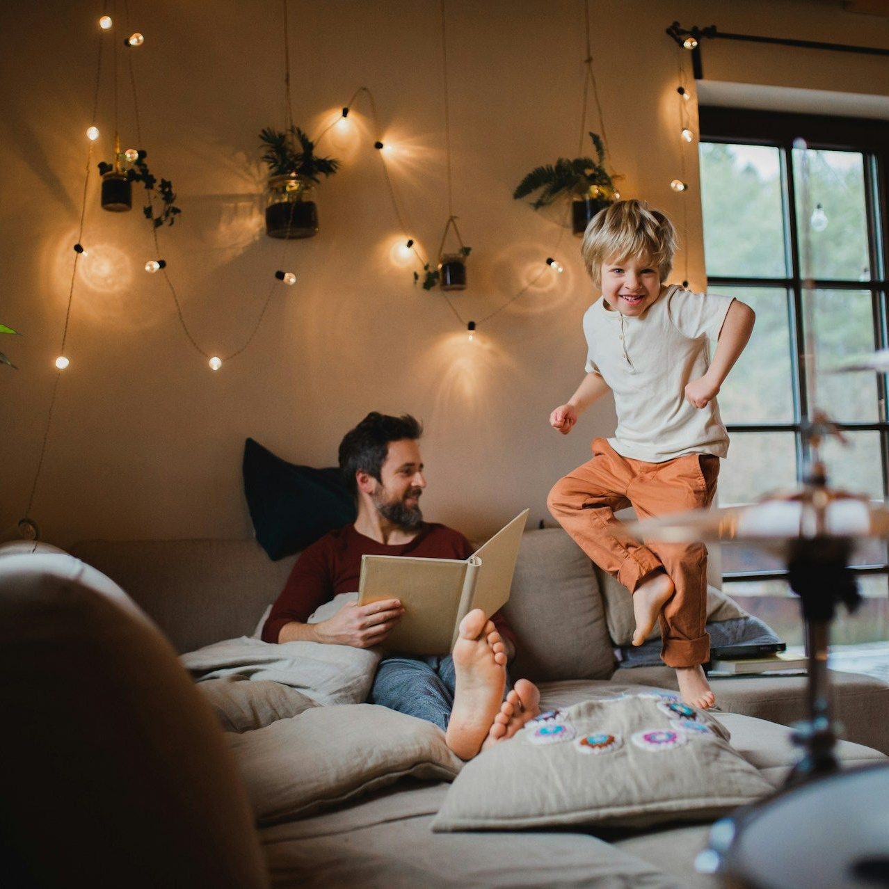 Man reading on couch as child jumps excitedly nearby, warm-toned living room, fairy lights.