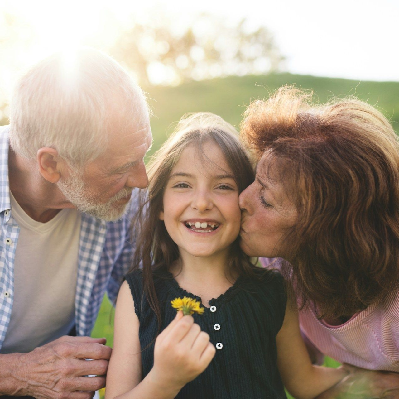 Grandparents smiling with a child outdoors; grandmother kisses the child's cheek.