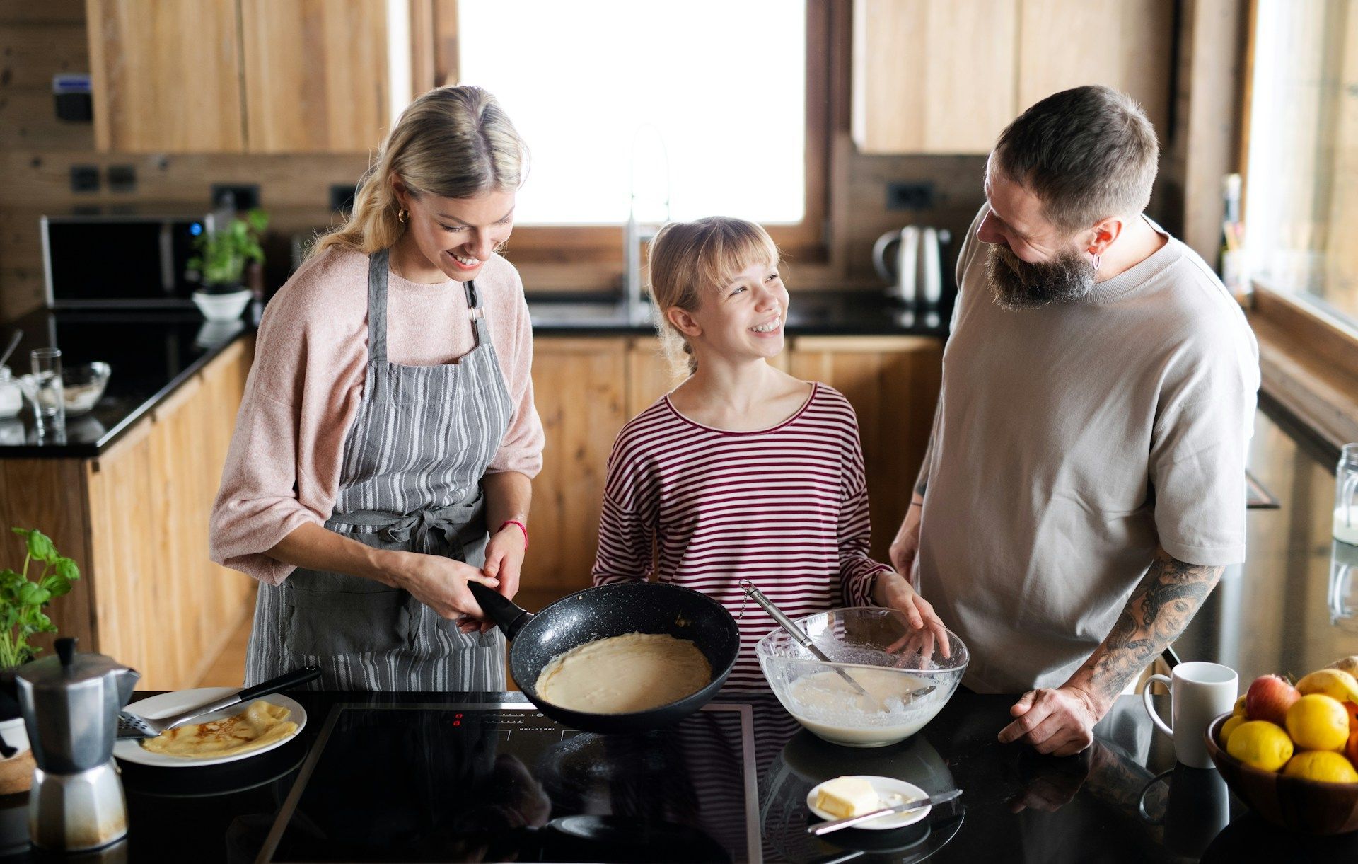 Family cooking pancakes in a modern kitchen: woman flips pancake, child smiles, man watches.