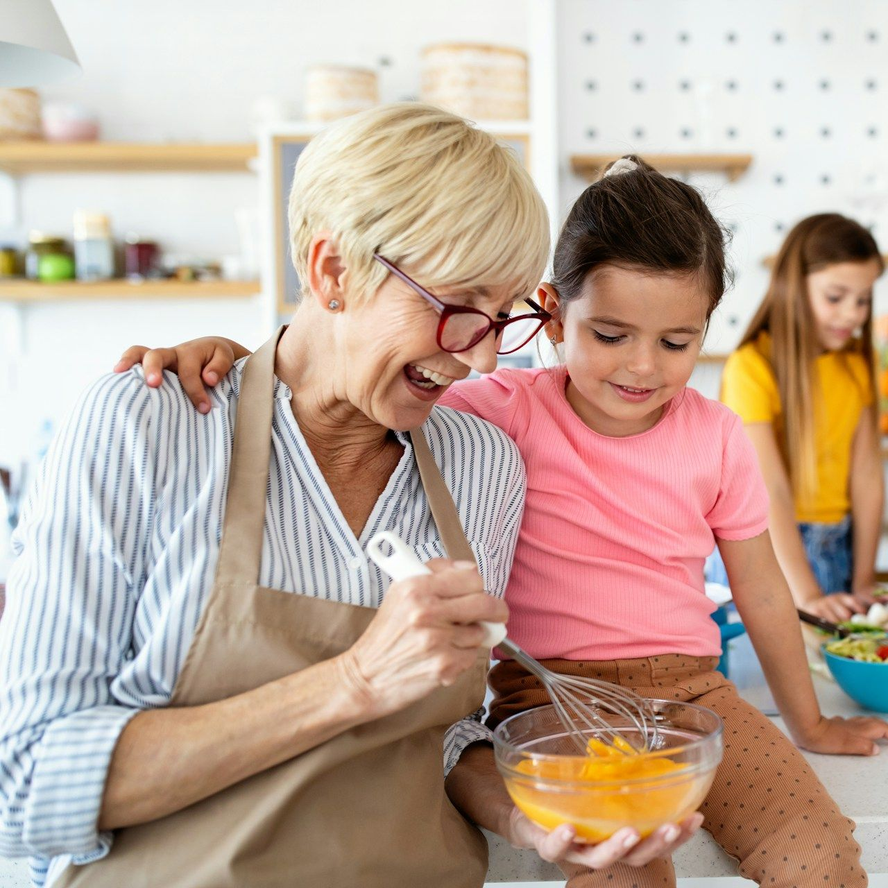 Woman with a child whisking ingredients in a kitchen, smiling. Another child in the background.