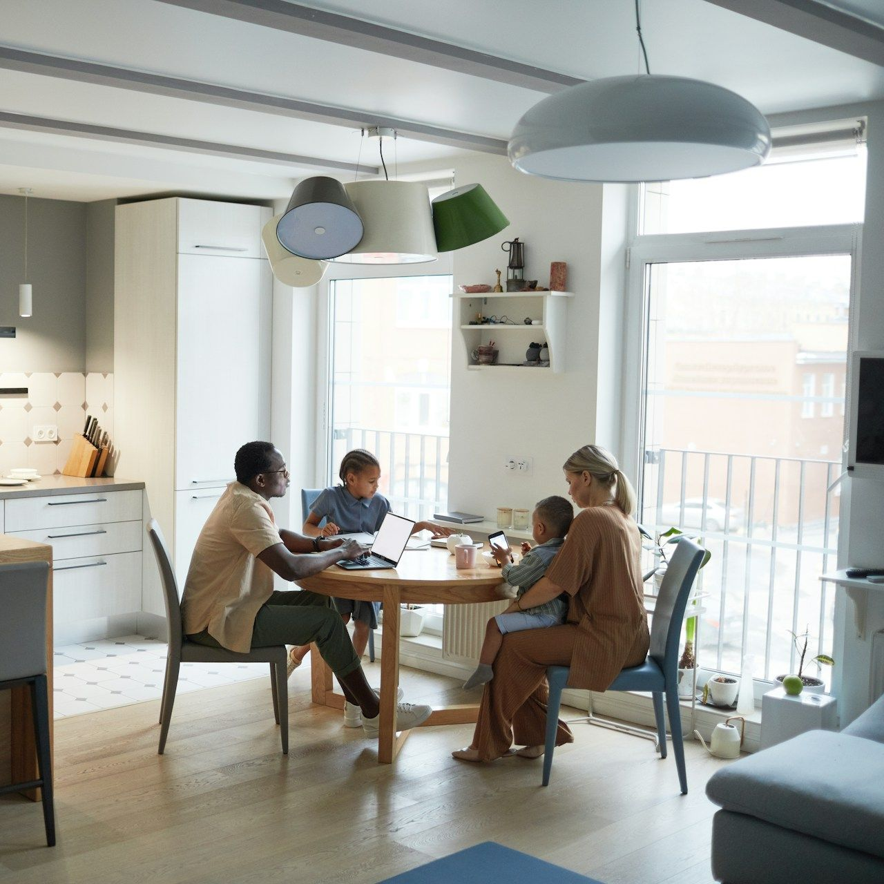 Family sits at a round table in a brightly lit room. A person holds a child while another works on a laptop.