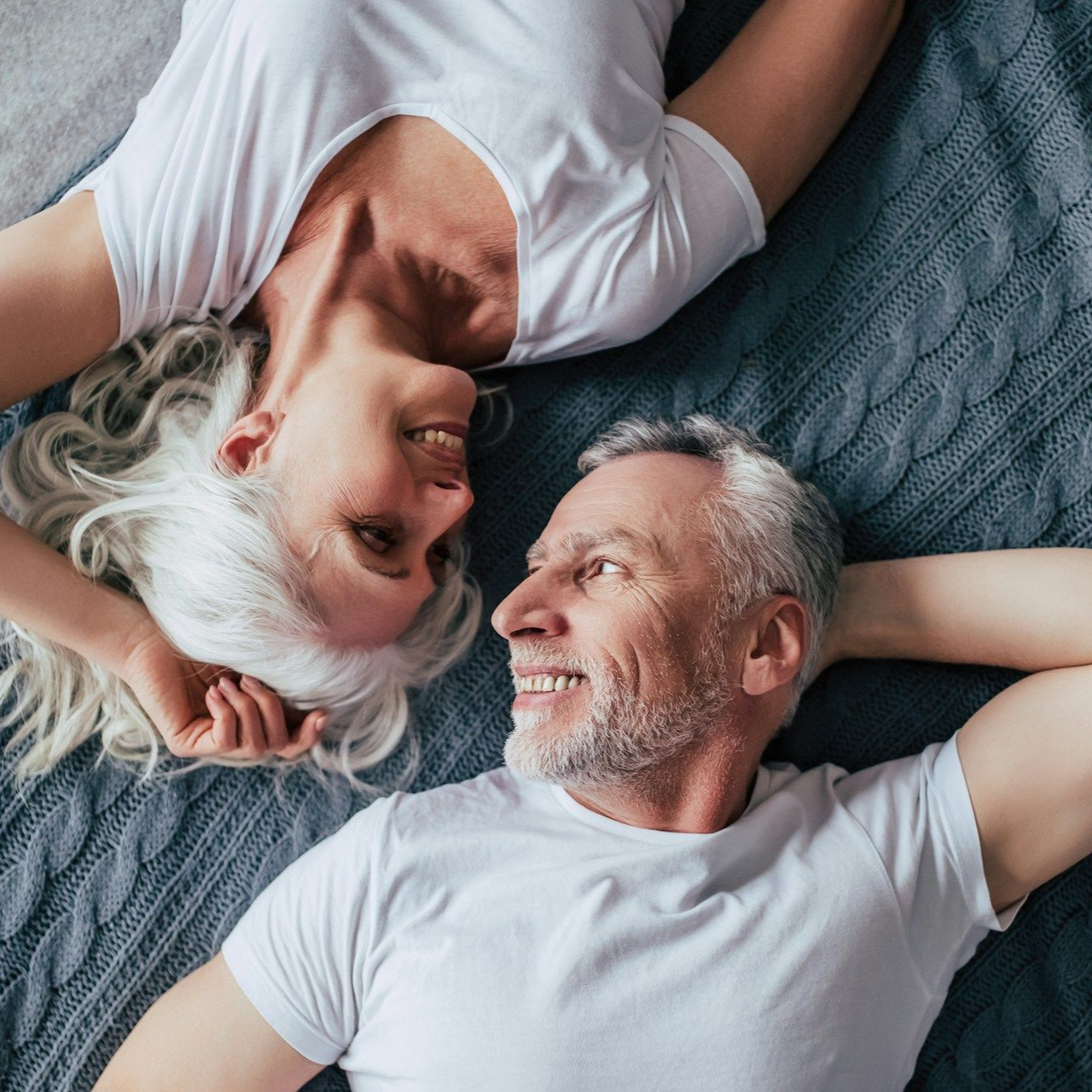 Smiling couple in white shirts, looking at each other while laying on a bed.