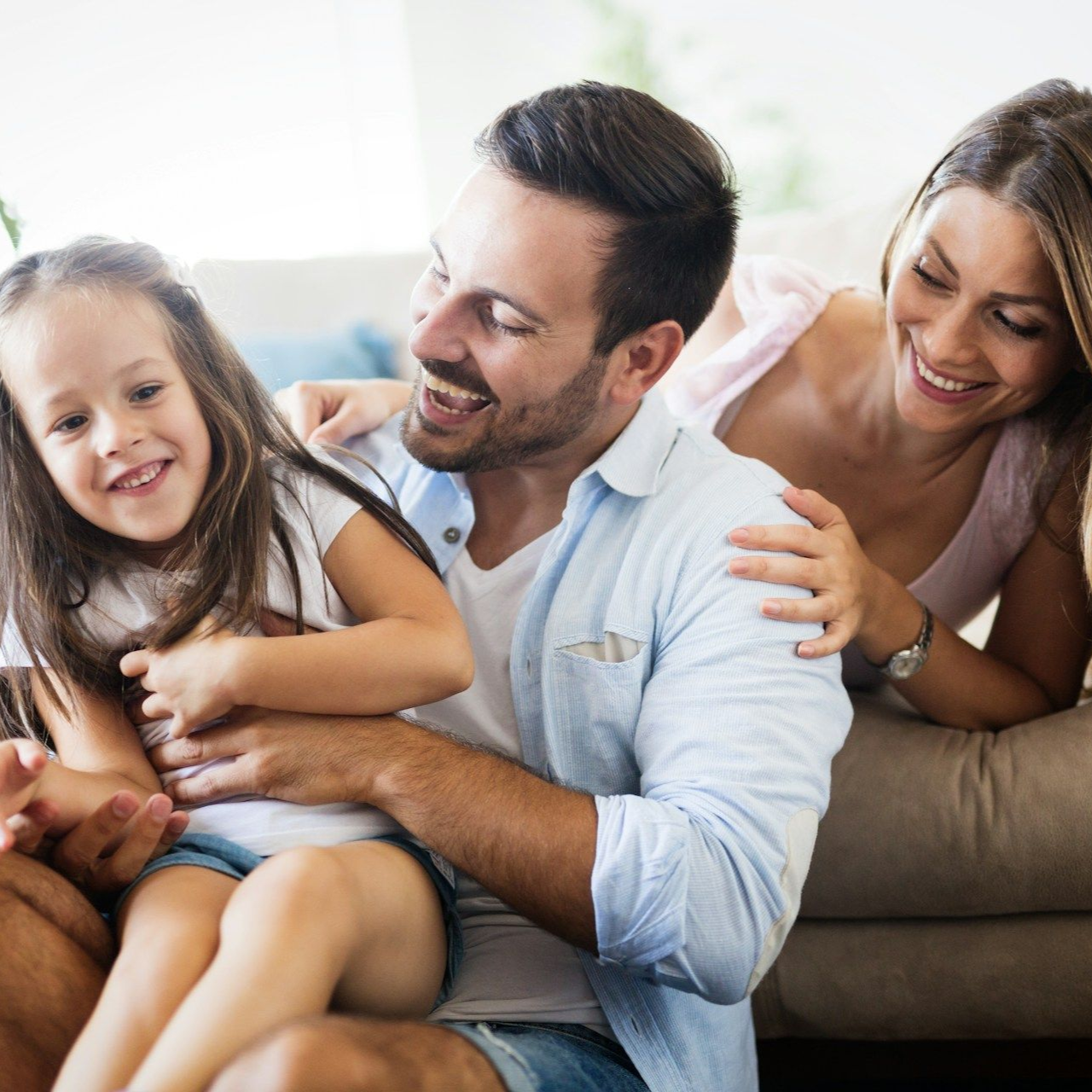 A family of three laughs together indoors. A father and daughter sit; a mother has her arms around them.