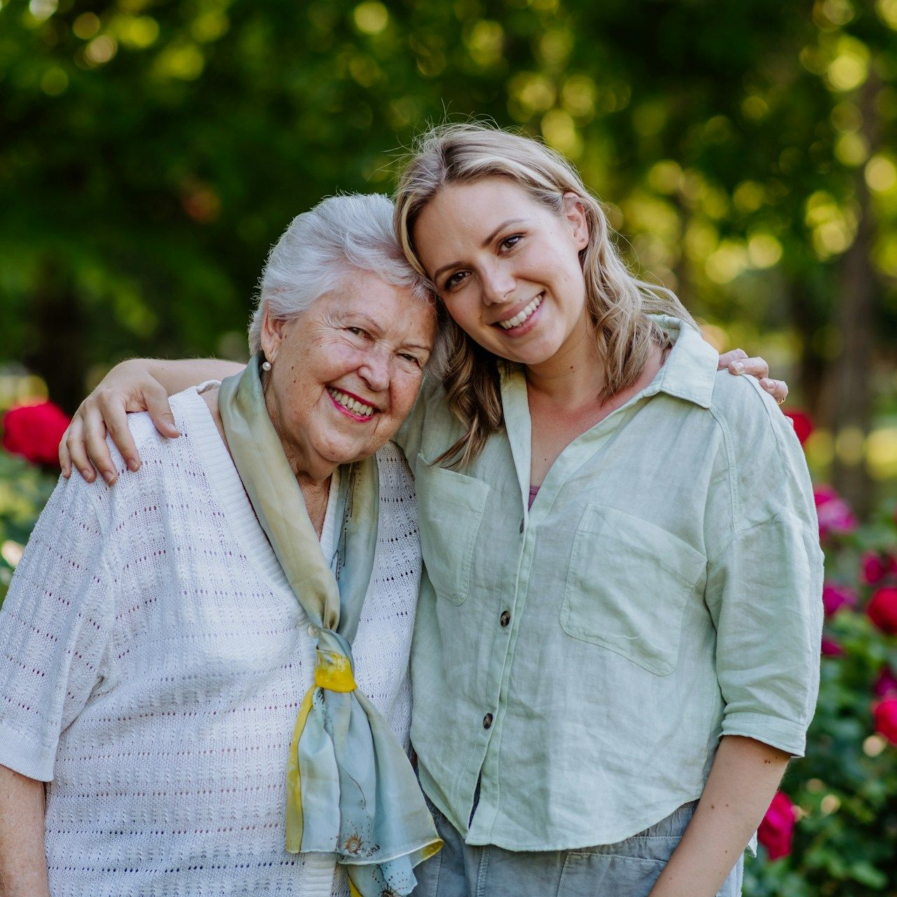 Woman with arm around elderly woman, smiling outdoors near roses.