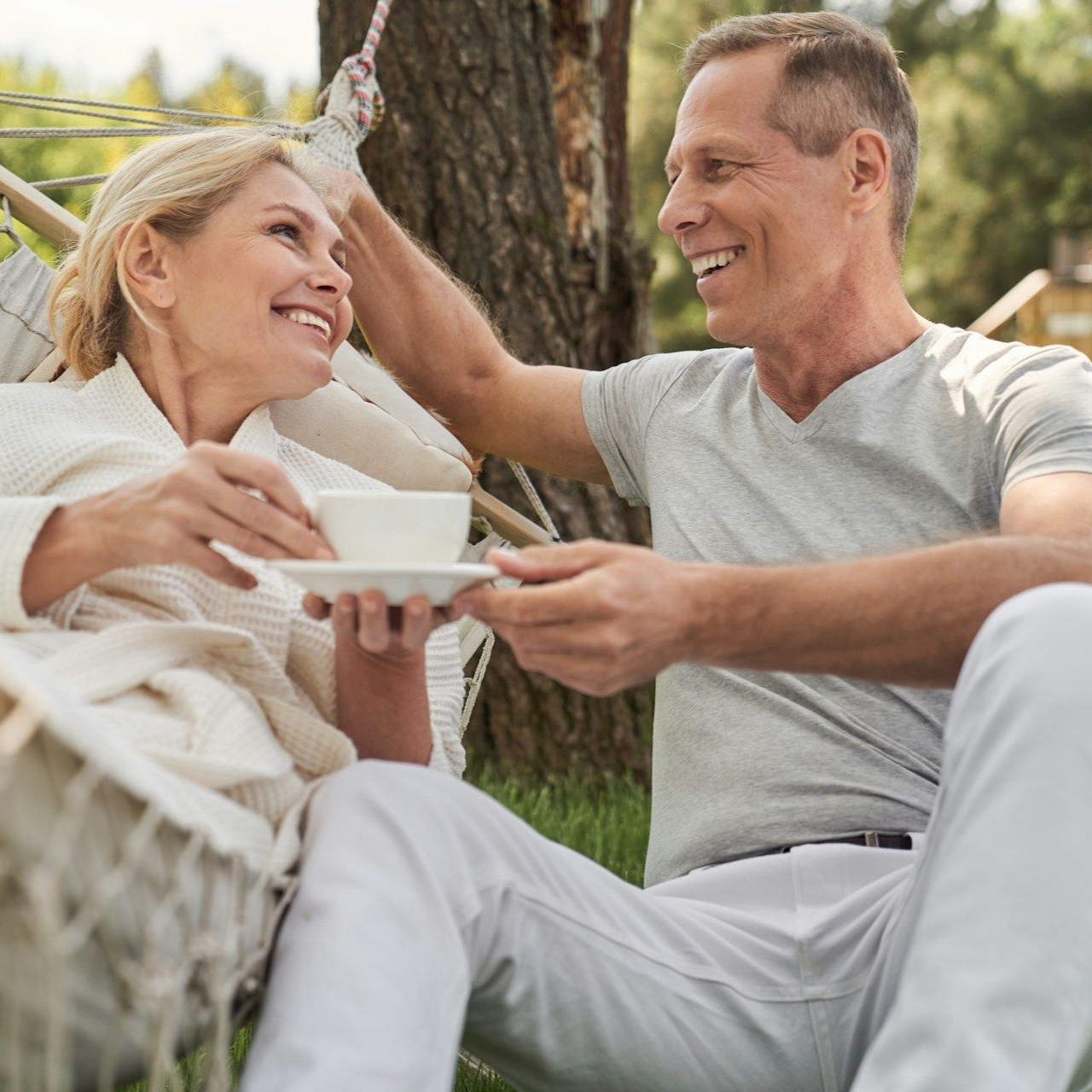 Couple in hammock, man smiling, touching woman's hair, she holds a teacup, outdoors.