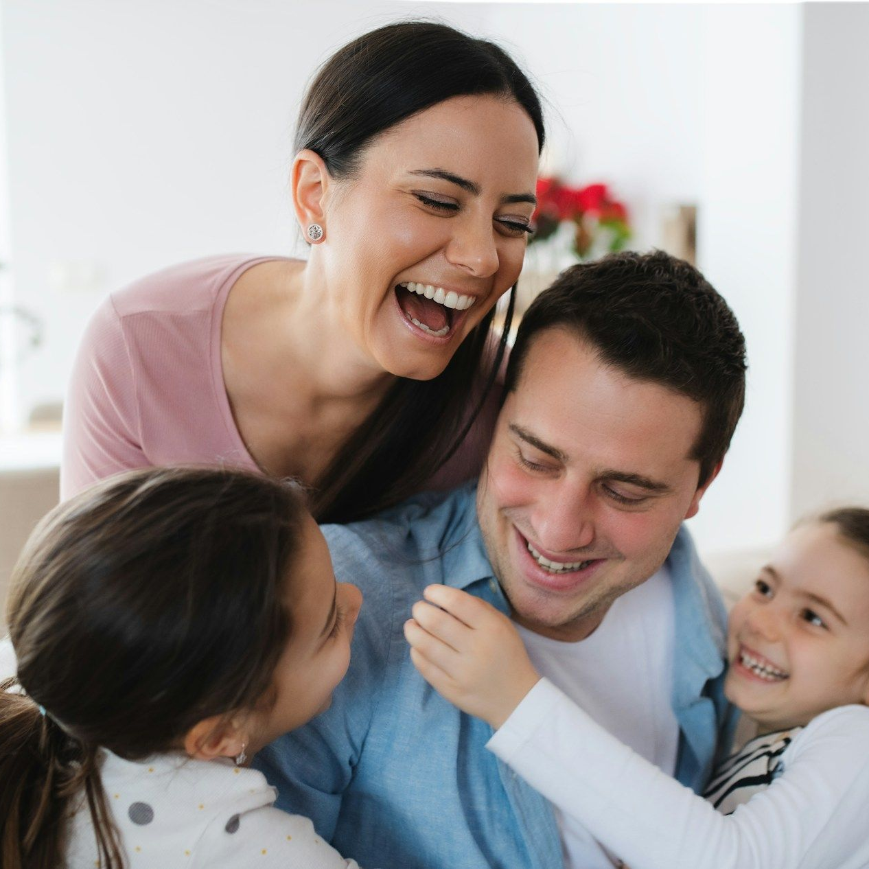 Family laughing and embracing in a well-lit room. Woman leans over man, two children hug.