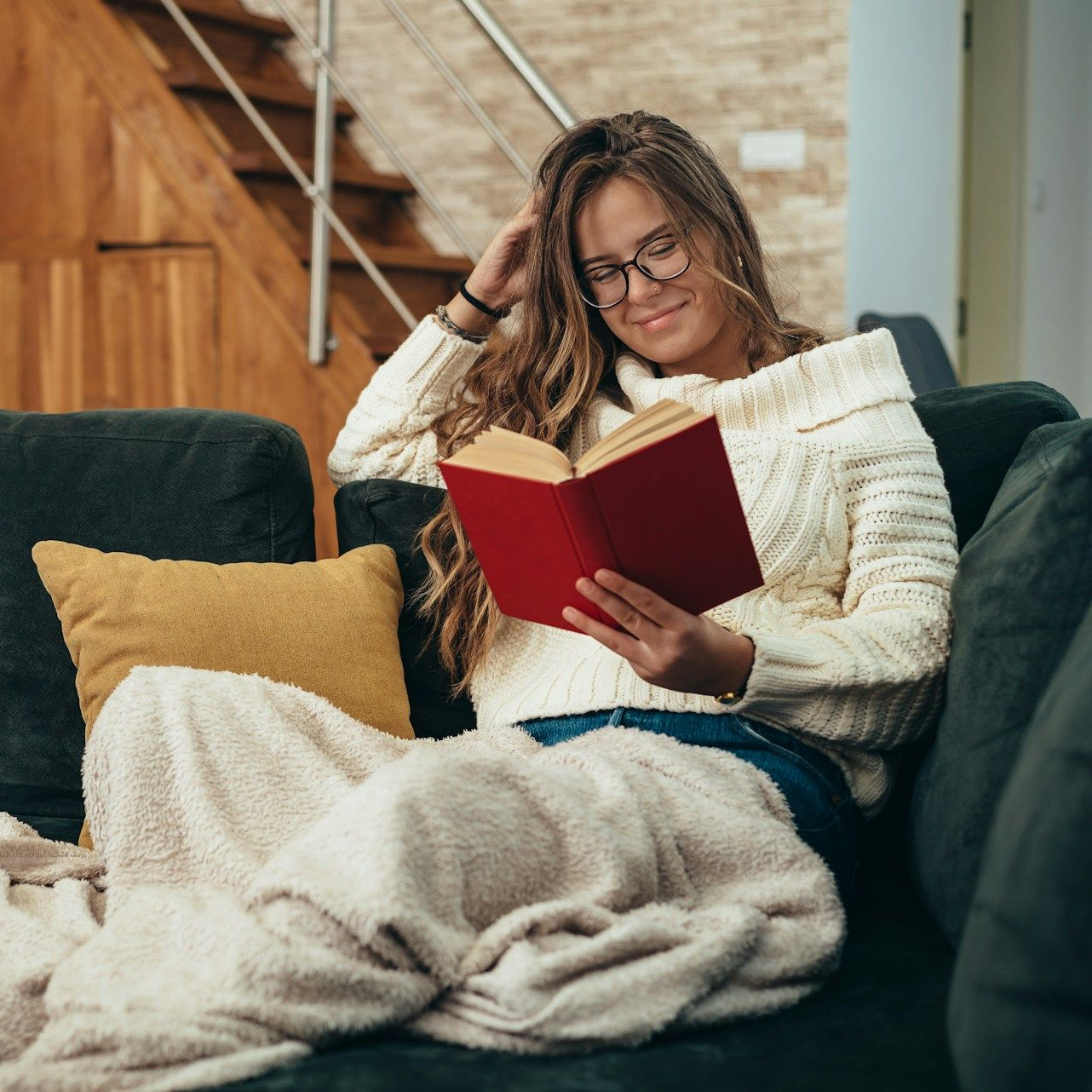 Woman wearing glasses, reading a red book on a couch with a blanket in a living room.