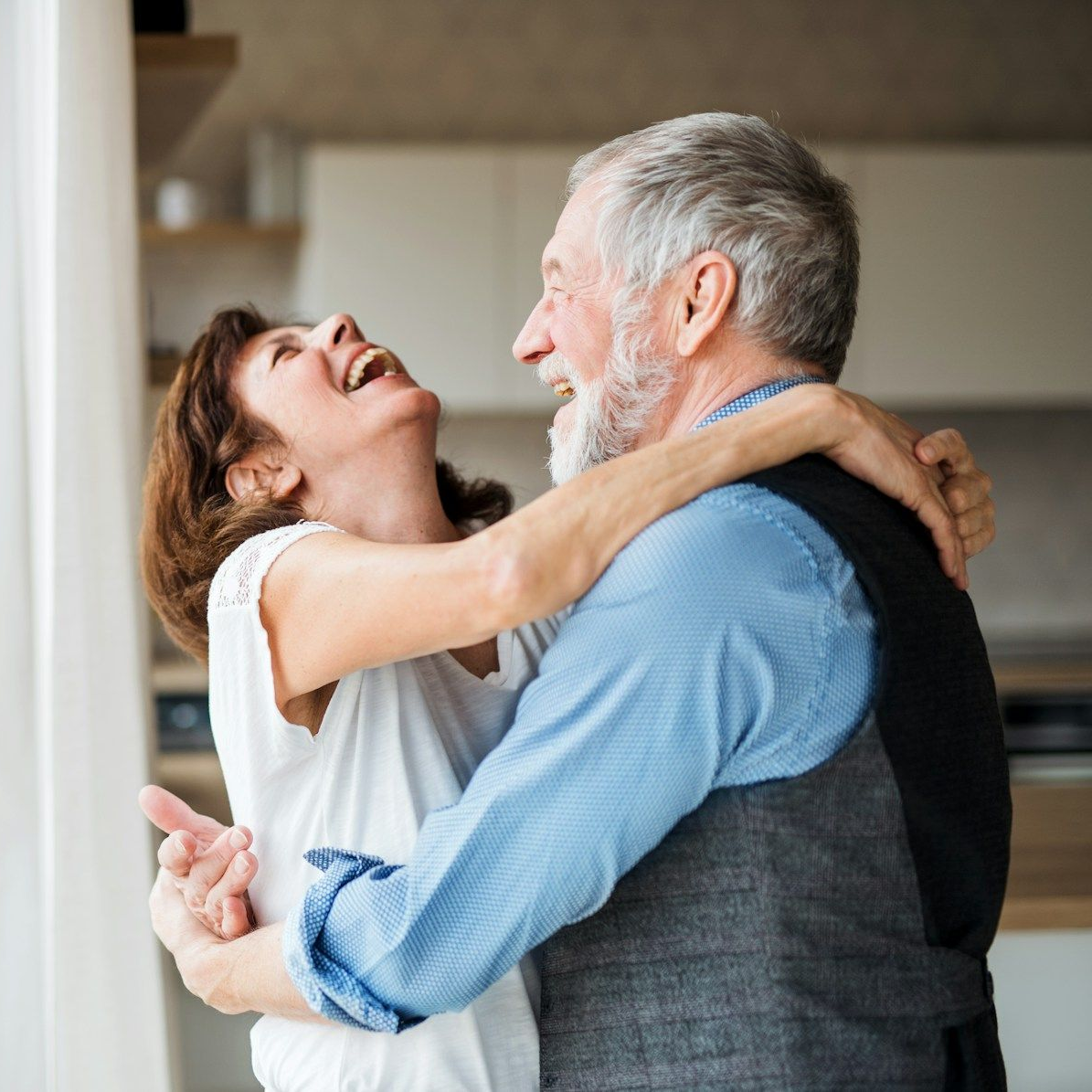 Older couple dancing and laughing indoors. Woman in white shirt, man in blue, gray vest.