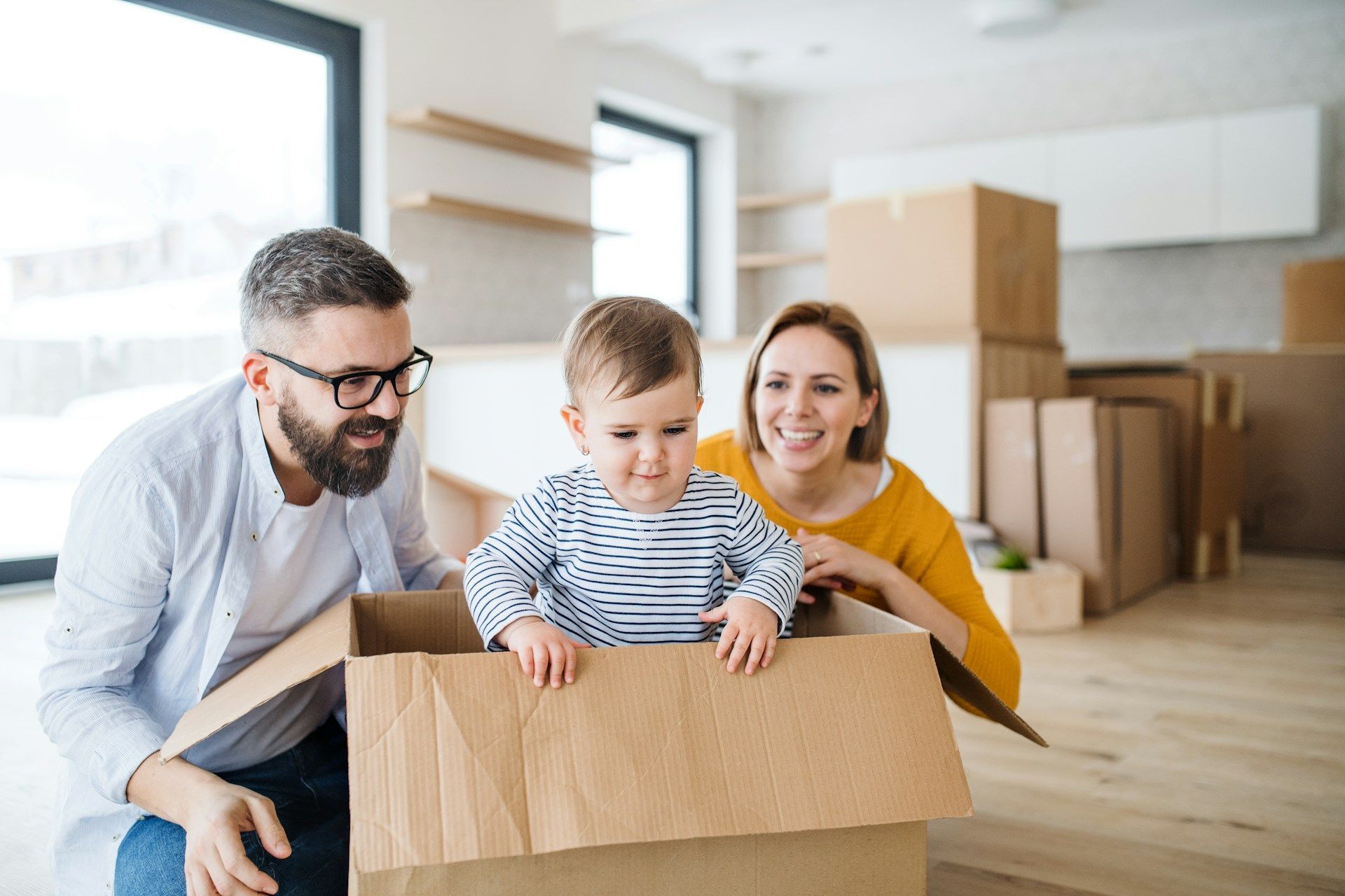 Family with a baby in a moving box, smiling in a new home. Cardboard boxes in the background.