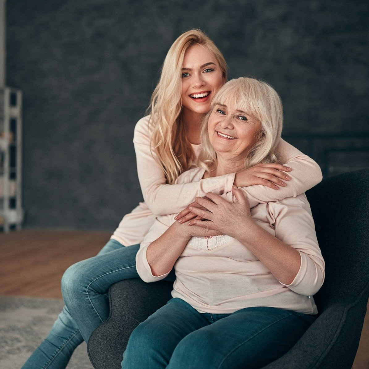 Young woman embraces older woman sitting in a chair, both smiling; indoor setting.