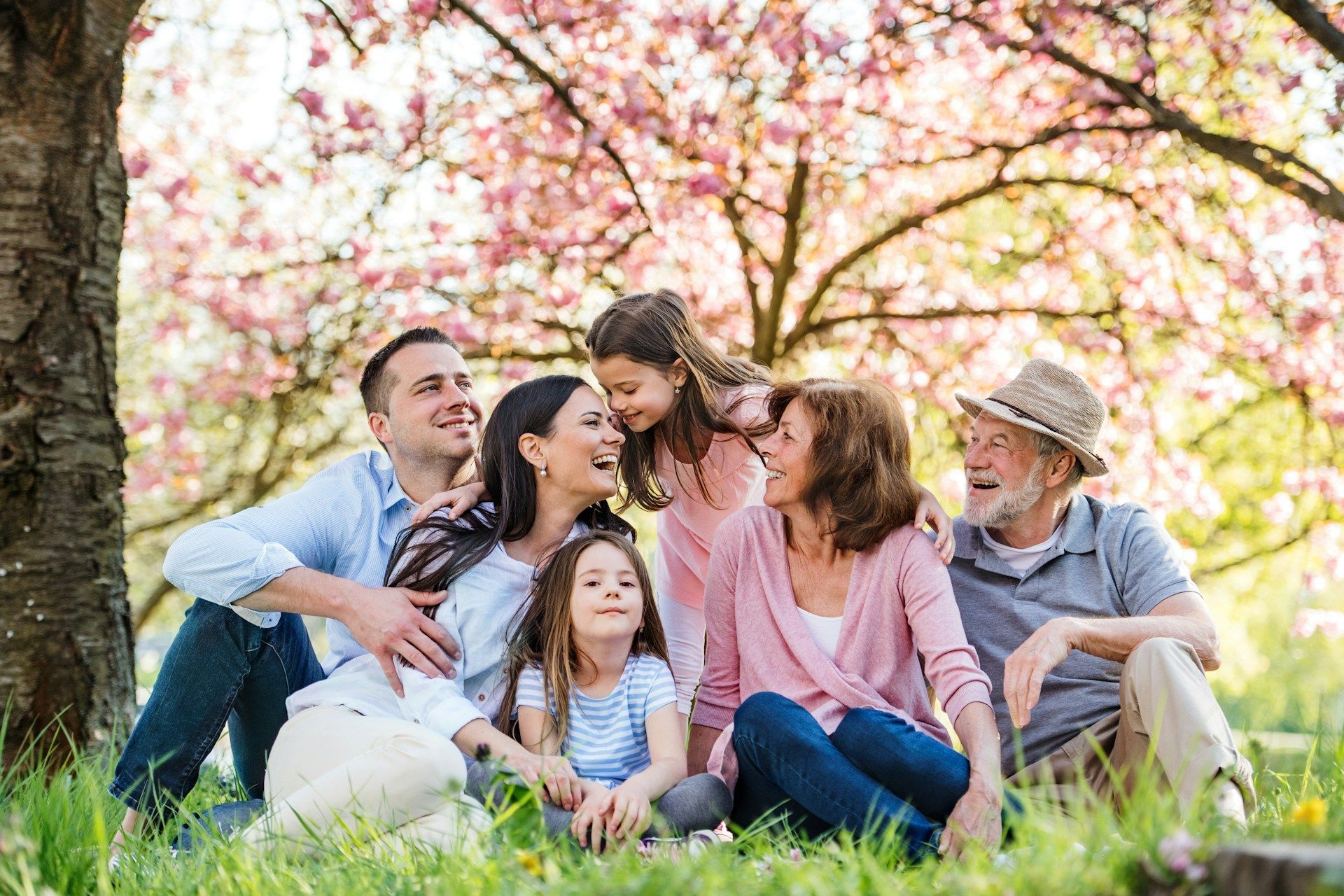 Family of six laughing together under a blooming pink tree in a sunny grassy field.