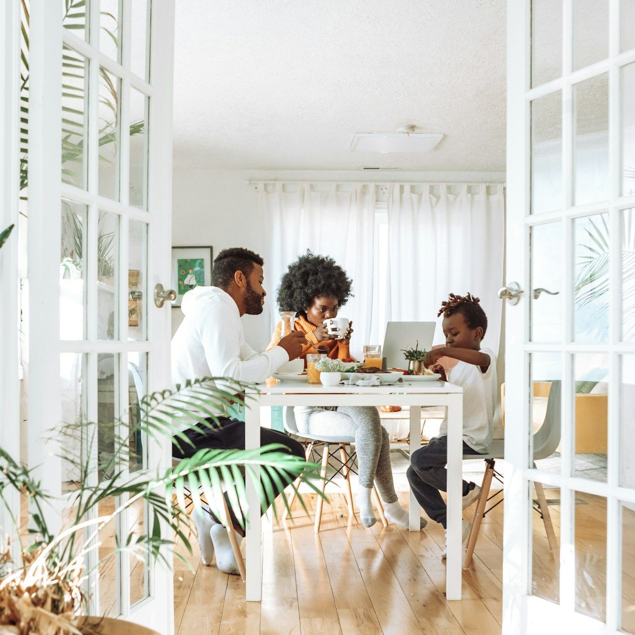 Family eating at a white table in a bright room, seen through French doors.