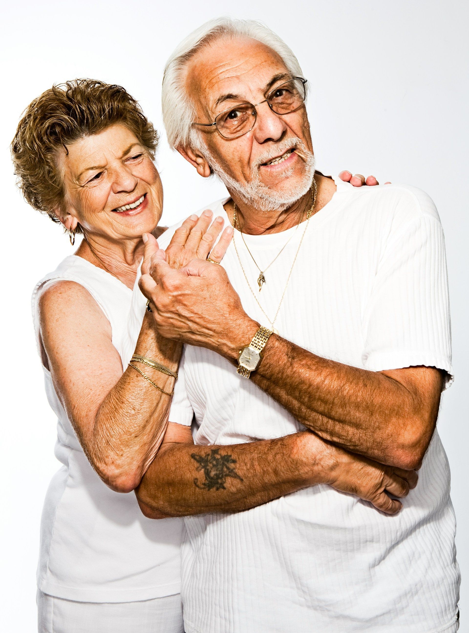 Elderly couple smiling, embracing. Woman's hand on man's hand. Man has arm tattoo, watch, glasses. 
