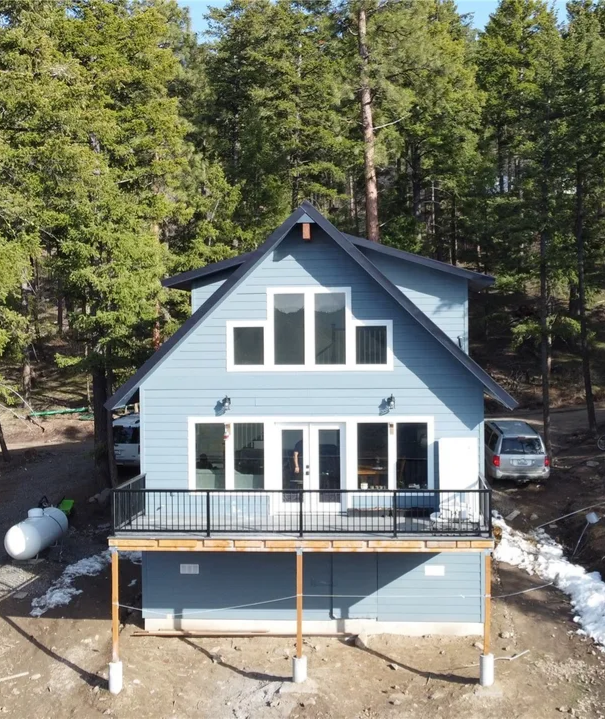Blue house with black roof and deck in a wooded area, gas tank on left, vehicle on right.