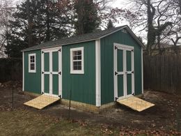 Tan shed with black roof, white trim, next to a building on a red-patterned walkway.