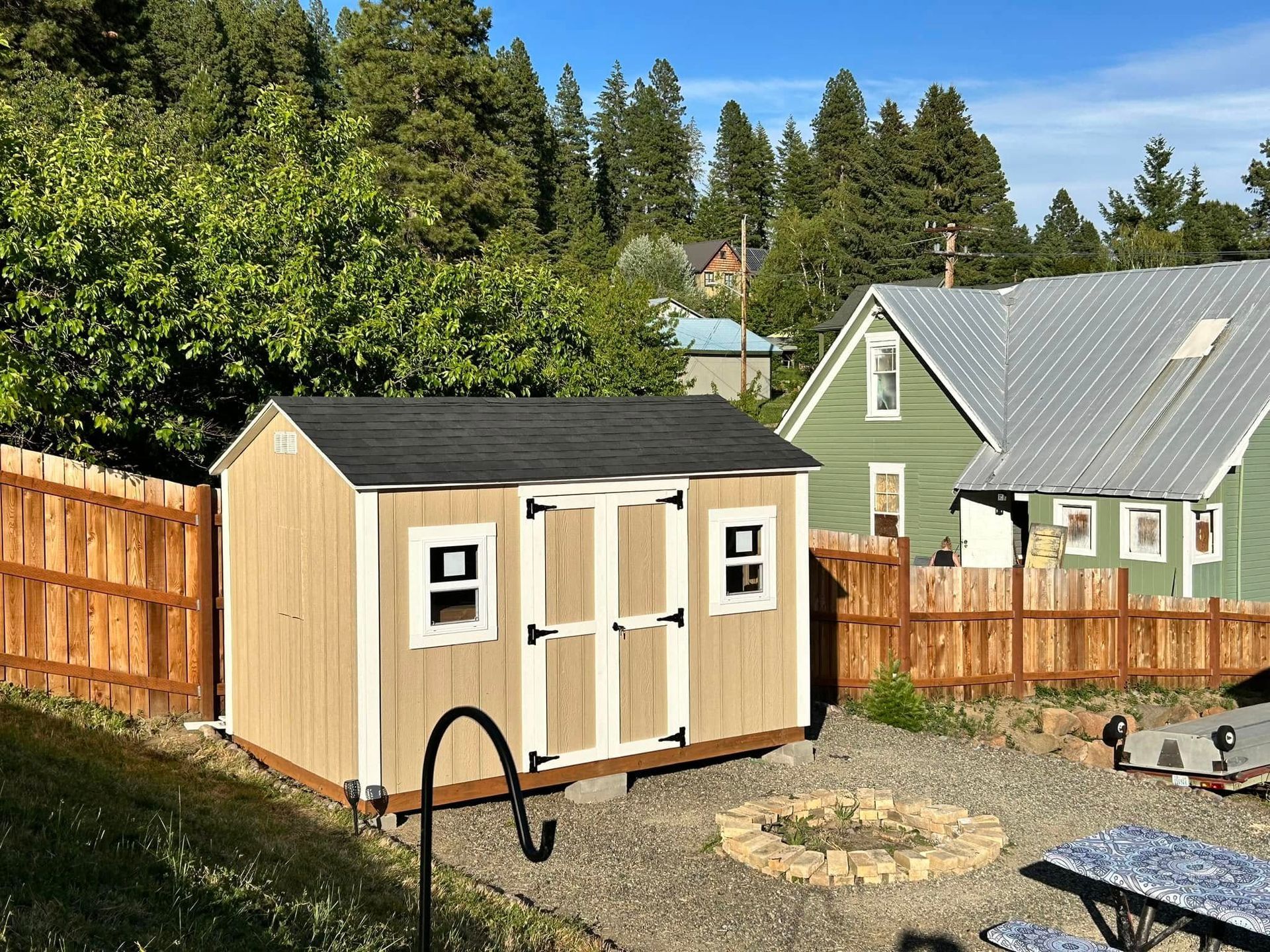 Tan shed with black roof in a backyard, brown wooden fence and green house visible, sunny day.