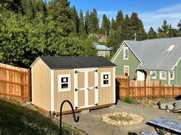 Tan shed with black roof in a backyard, brown wooden fence and green house visible, sunny day.
