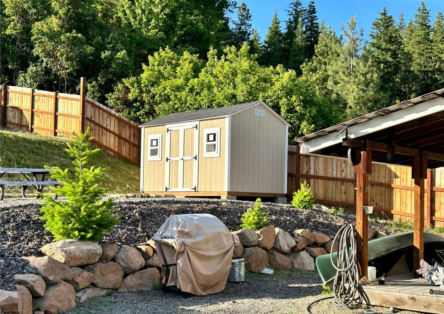A gray shed with a white door sits on a concrete pad. A white fence and trees are visible in the background.