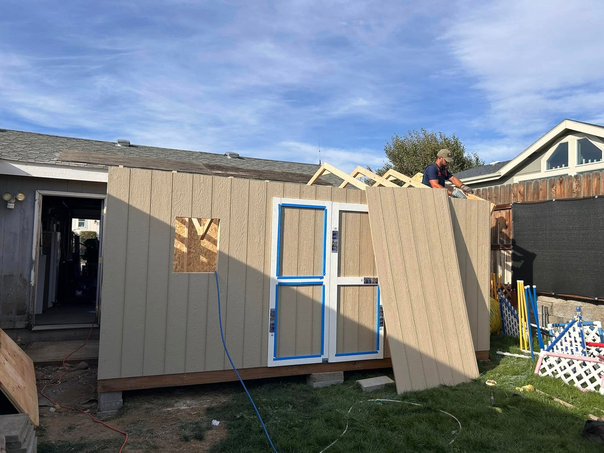 Construction of a shed in a backyard. Man on roof, attaching panel to side with door and window.