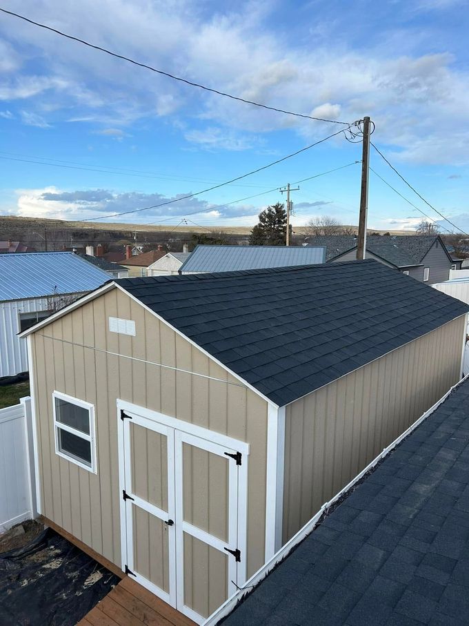 Tan shed with black roof and white trim under a partly cloudy sky.