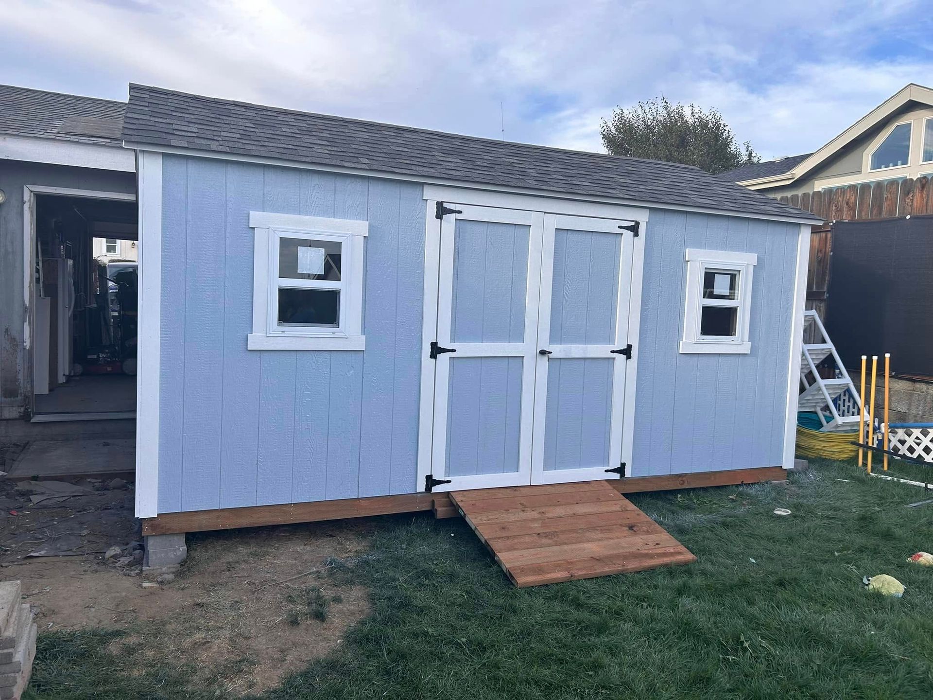 A gray shed with a white door sits on a concrete pad. A white fence and trees are visible in the background.