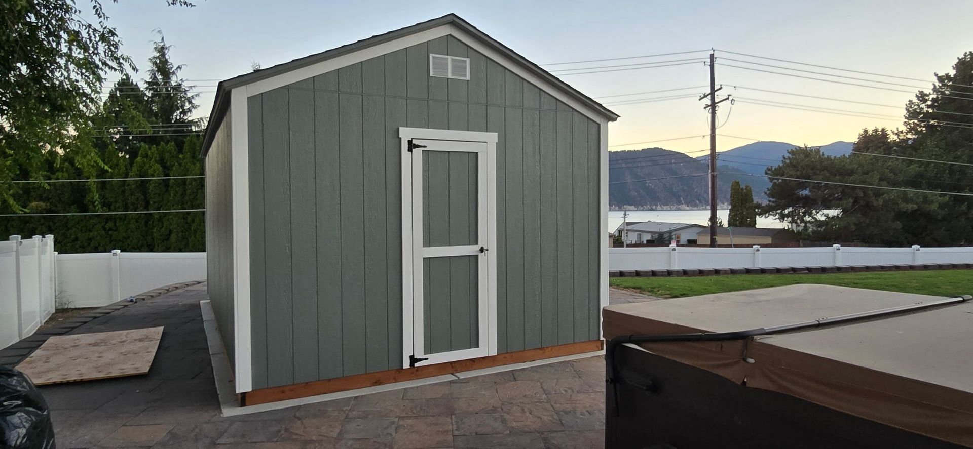 A gray shed with a white door sits on a concrete pad. A white fence and trees are visible in the background.
