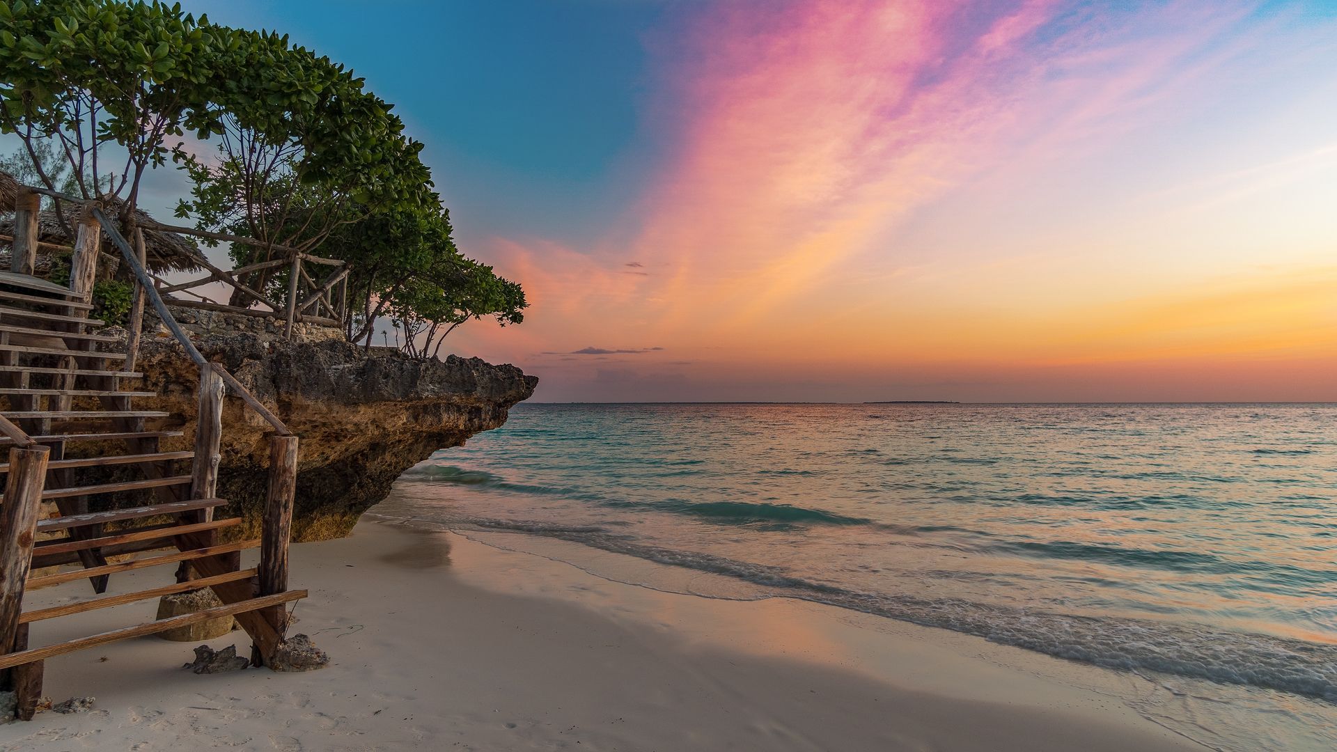 A wooden staircase leading up to a rock formation on a beach at sunset.