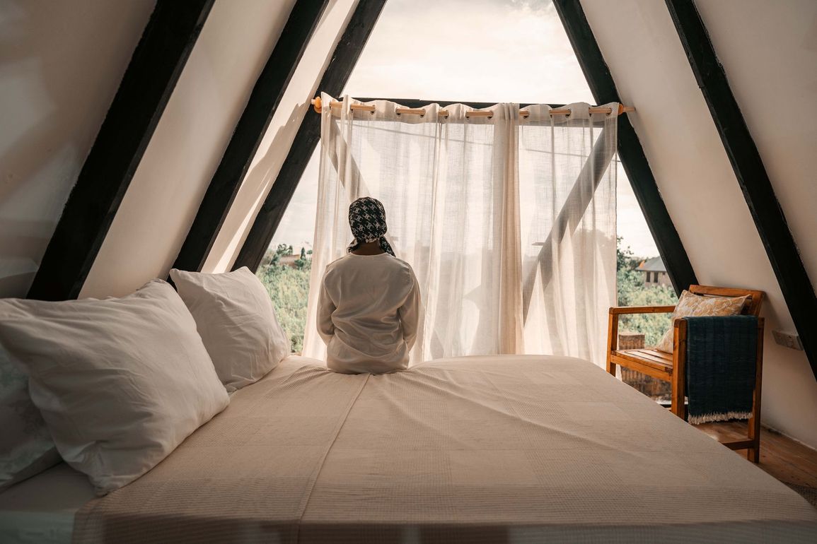 Person sitting on a bed in a bright A-frame cabin, facing a sheer-curtained window.