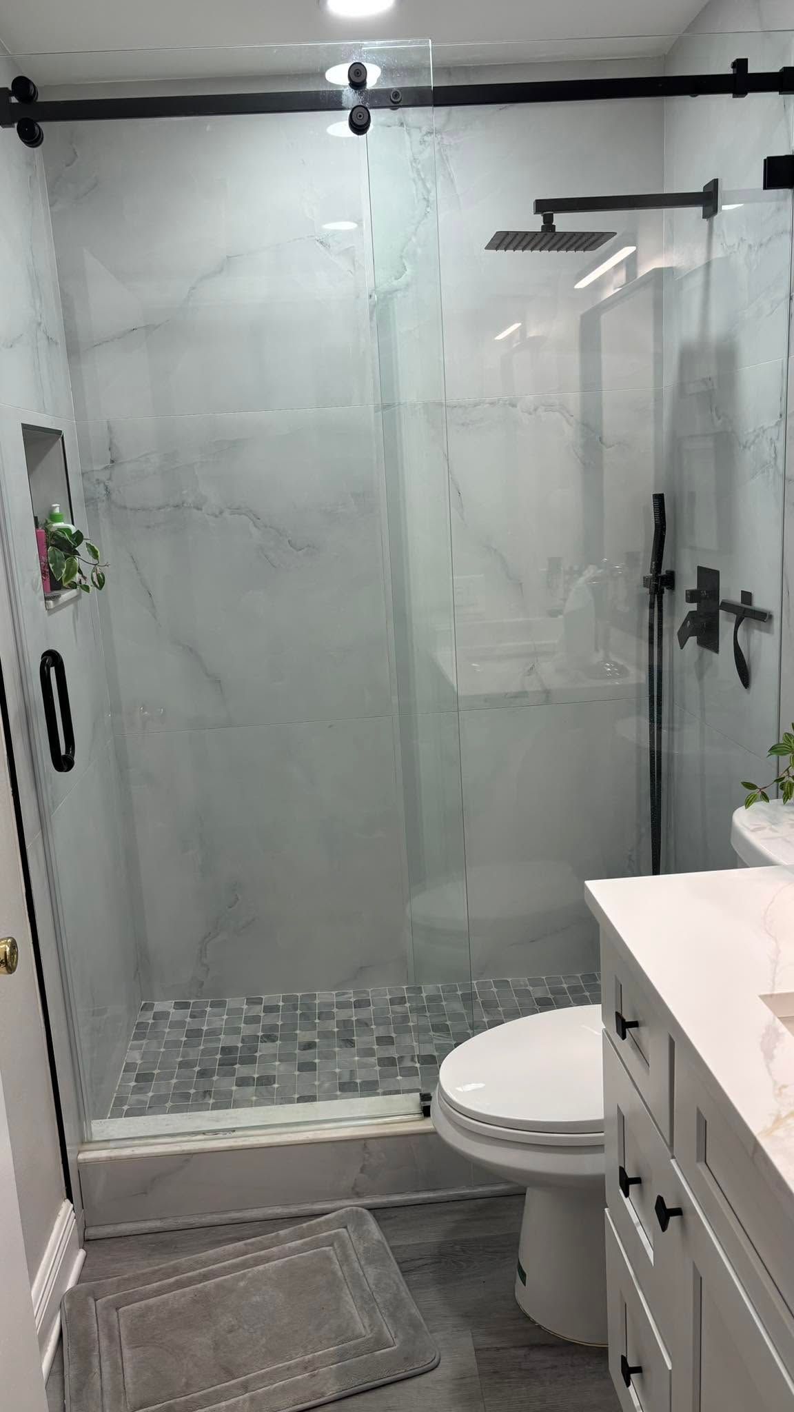 Small bathroom with glass shower, toilet, gray floor mat, and white vanity.