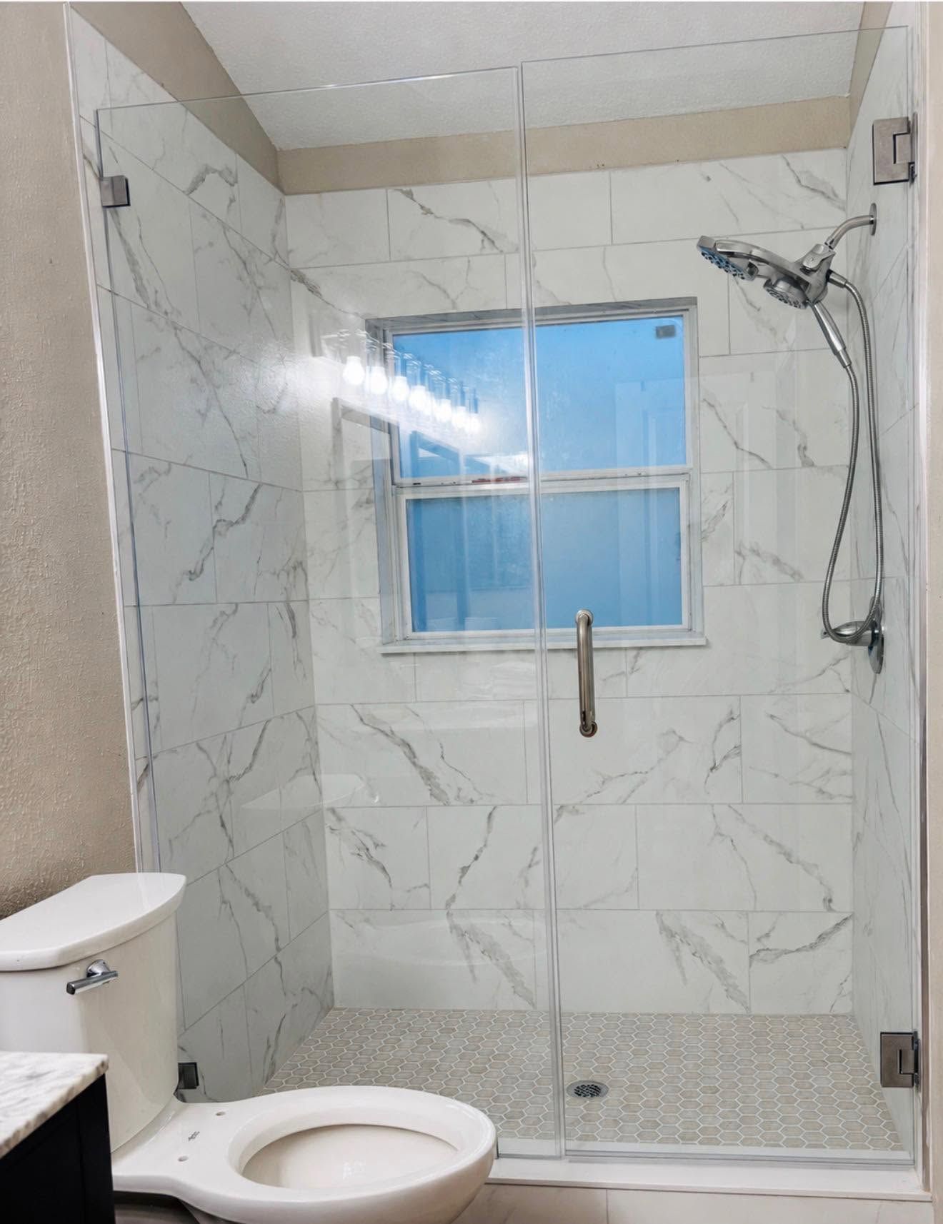 Modern marble-tiled bathroom shower with glass door, window, toilet, and chrome fixtures