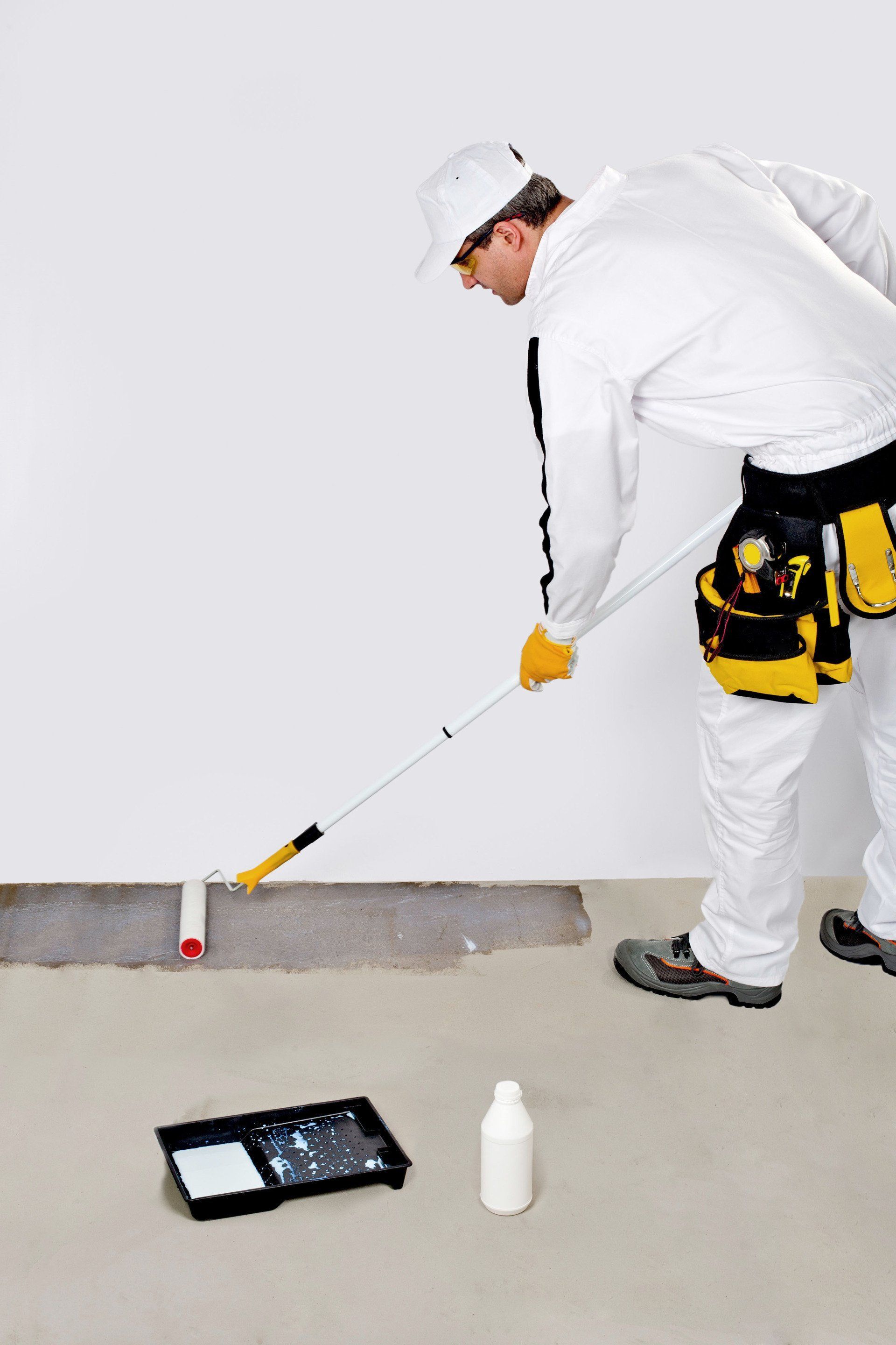 Painter in white overalls rolling paint on a concrete floor, with tools, paint tray, and bottle nearby.