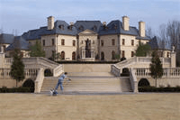 Man and dog walk on stairs leading to a large, beige mansion with blue roof.