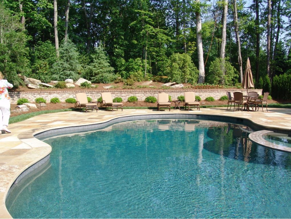 Swimming pool with chaise lounges, a stone patio, and a brick retaining wall with trees in the background.