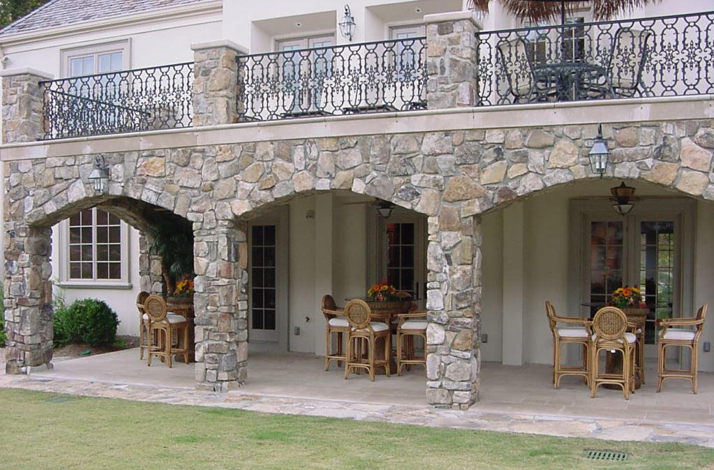 Stone archway with patio furniture, supporting a balcony on a light-colored building.