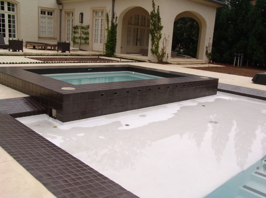 An outdoor pool with a raised, square hot tub. The tub is dark-tiled. White pool surface. Light beige patio, a white building.