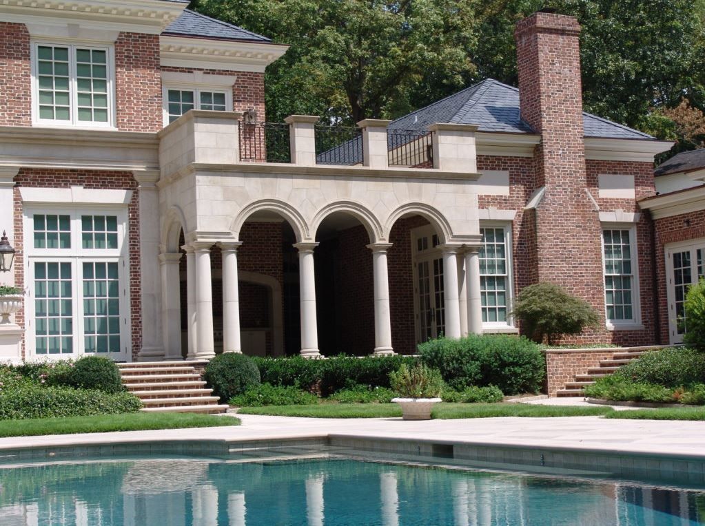 Brick mansion with a pool. Features arched columns, a balcony, and lush greenery.