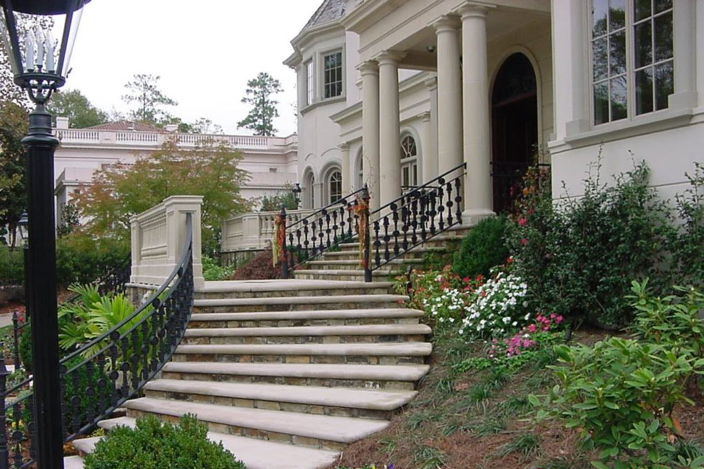 Stone steps lead to a large white mansion with columns, surrounded by flowers and greenery.