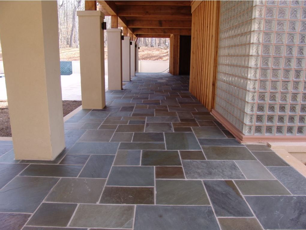 Stone tile walkway under a building with columns and a glass block wall.