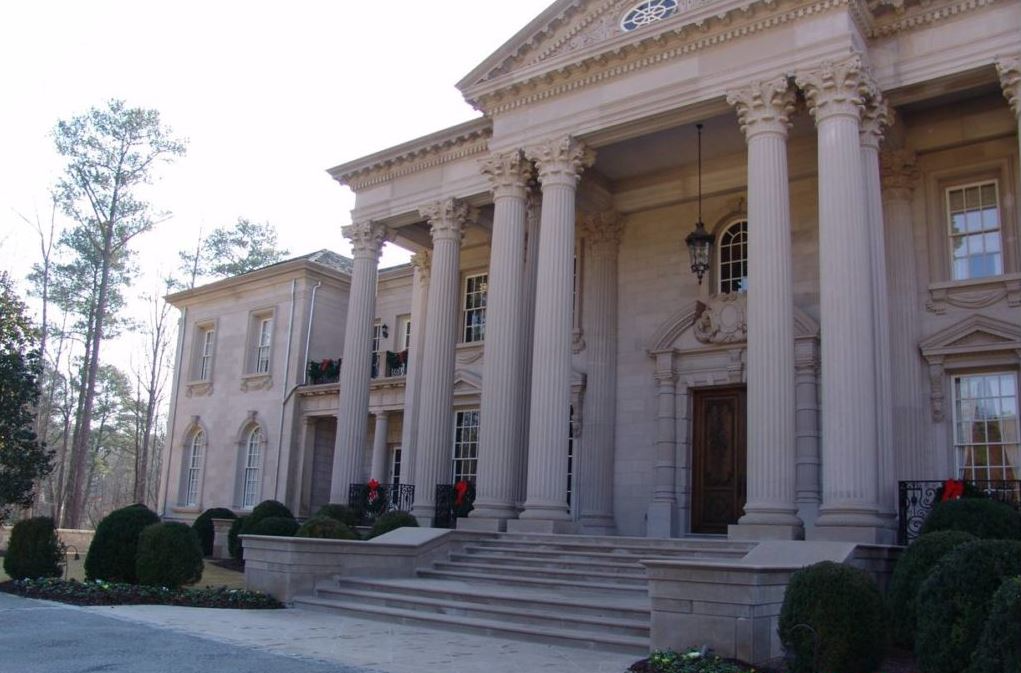 A large, light-colored mansion with columns and steps; surrounded by shrubbery and trees.