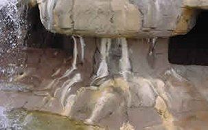 Water flows from a rock formation, light brown with some white streaks and a dark interior.
