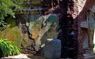 Stone pillar in front of a weathered mural on a brick wall with foliage.