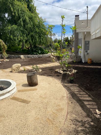 Gravel backyard with a fire pit, stone bench, trees, and a house beside a dirt garden bed