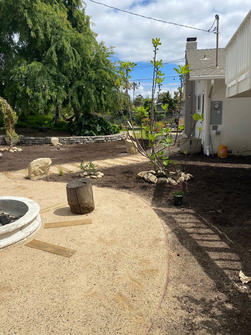 Gravel backyard with a fire pit, stone bench, trees, and a house beside a dirt garden bed
