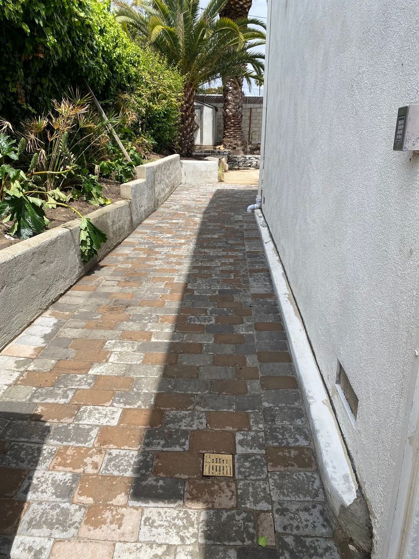Narrow brick walkway beside a white wall, bordered by lush greenery, leading to a gate.
