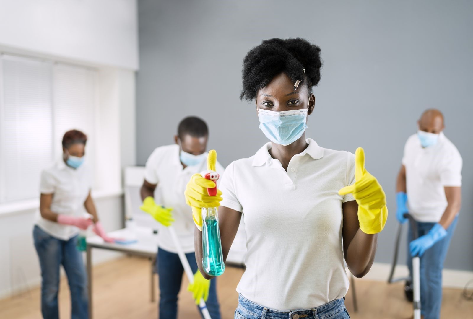 A woman wearing a mask is holding a spray bottle and giving a thumbs up.