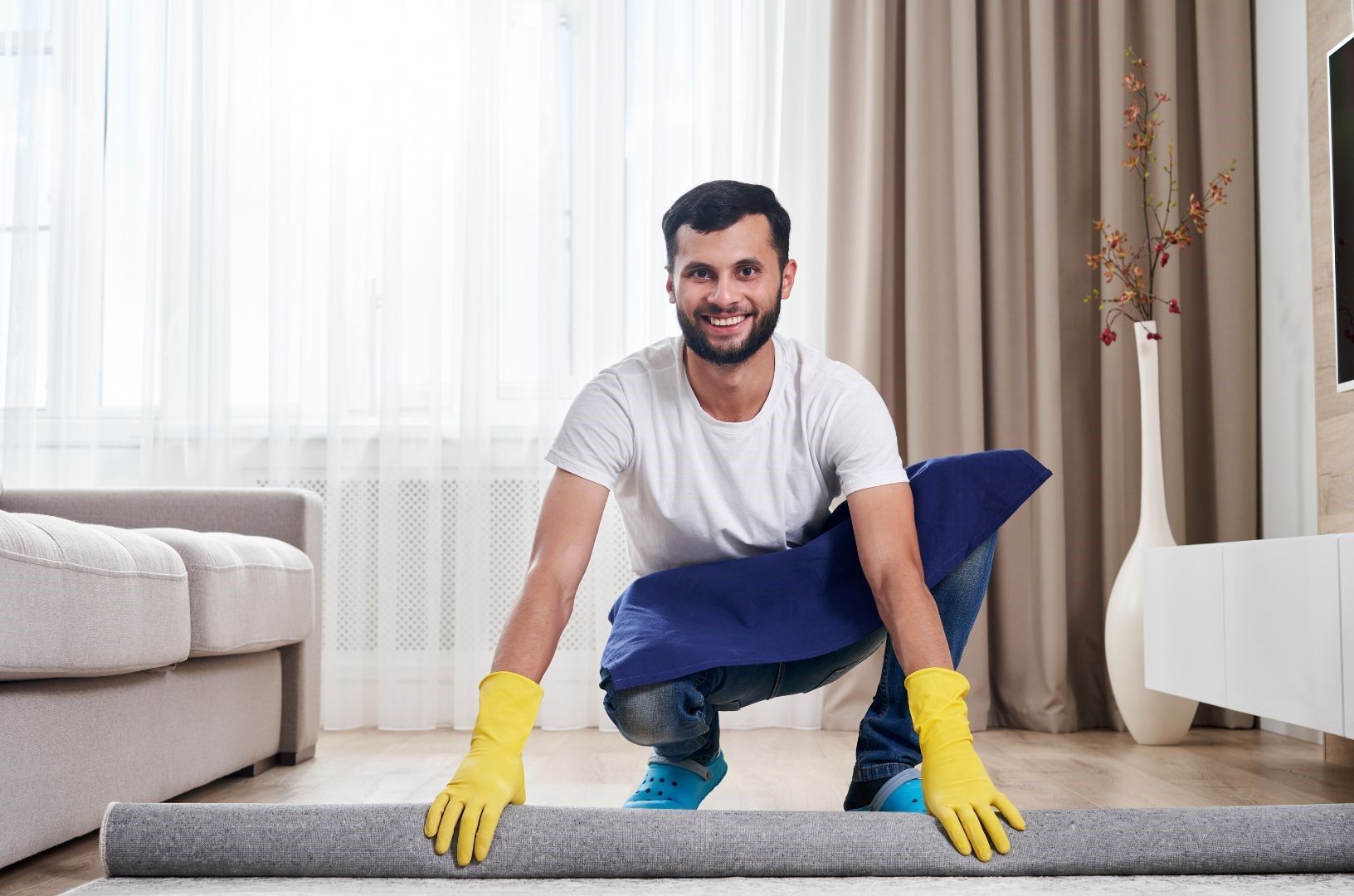A man wearing yellow gloves is cleaning a carpet in a living room.